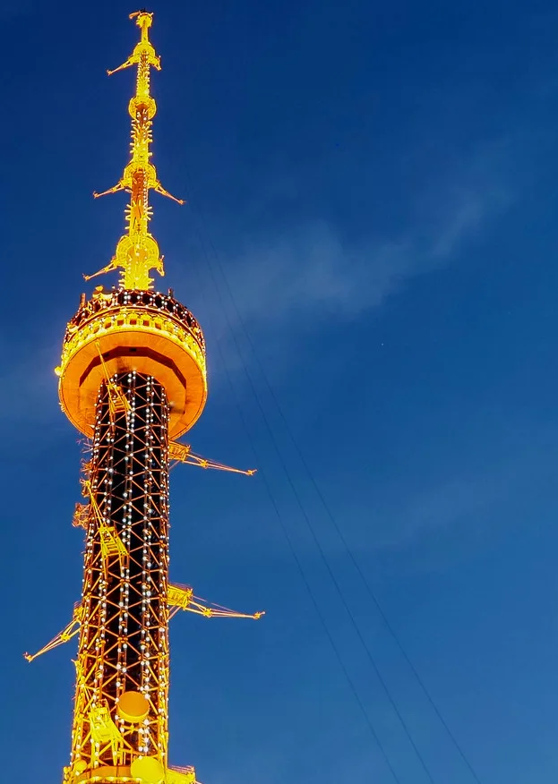 A low-angle shot of a tall, ornate, golden-lit tower against a clear blue sky, featuring decorative elements and lights. The…