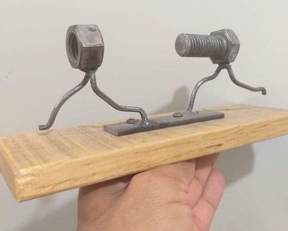 A hand holds a small wooden plank with a metal contraption attached: one side is a hex nut, the other a bolt, both mounted…