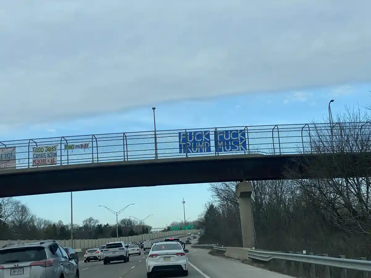 A highway scene with cars driving under an overpass where multiple protest signs are taped to the railing. Two large blue…