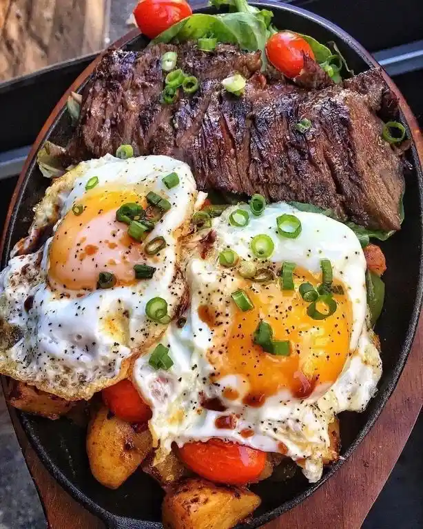A close-up photo of a hearty meal in a black cast iron skillet, featuring two sunny-side-up eggs with runny yolks, a large…