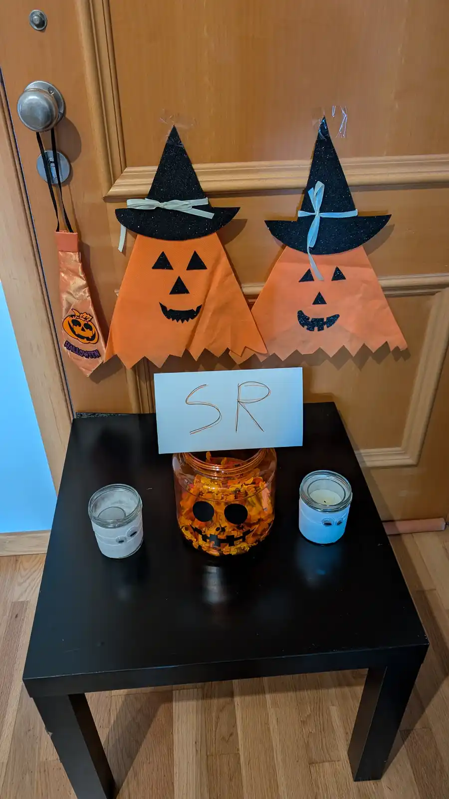 A Halloween candy station set up on a black table in front of a wooden door, featuring two orange paper jack-o'-lantern…