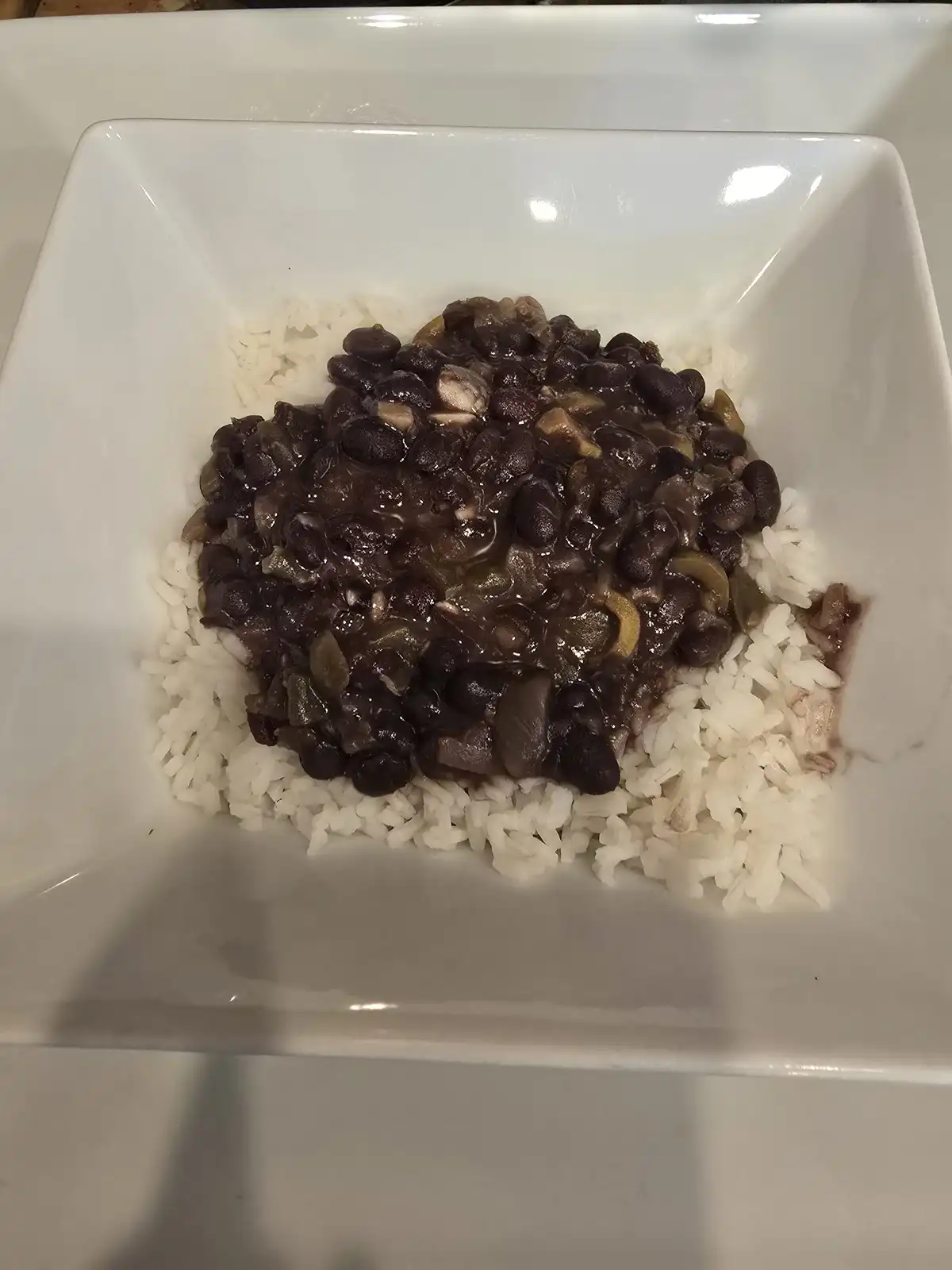 A white square bowl contains a serving of white rice topped with a dark, glossy black bean stew. The text overlay reads 'i…