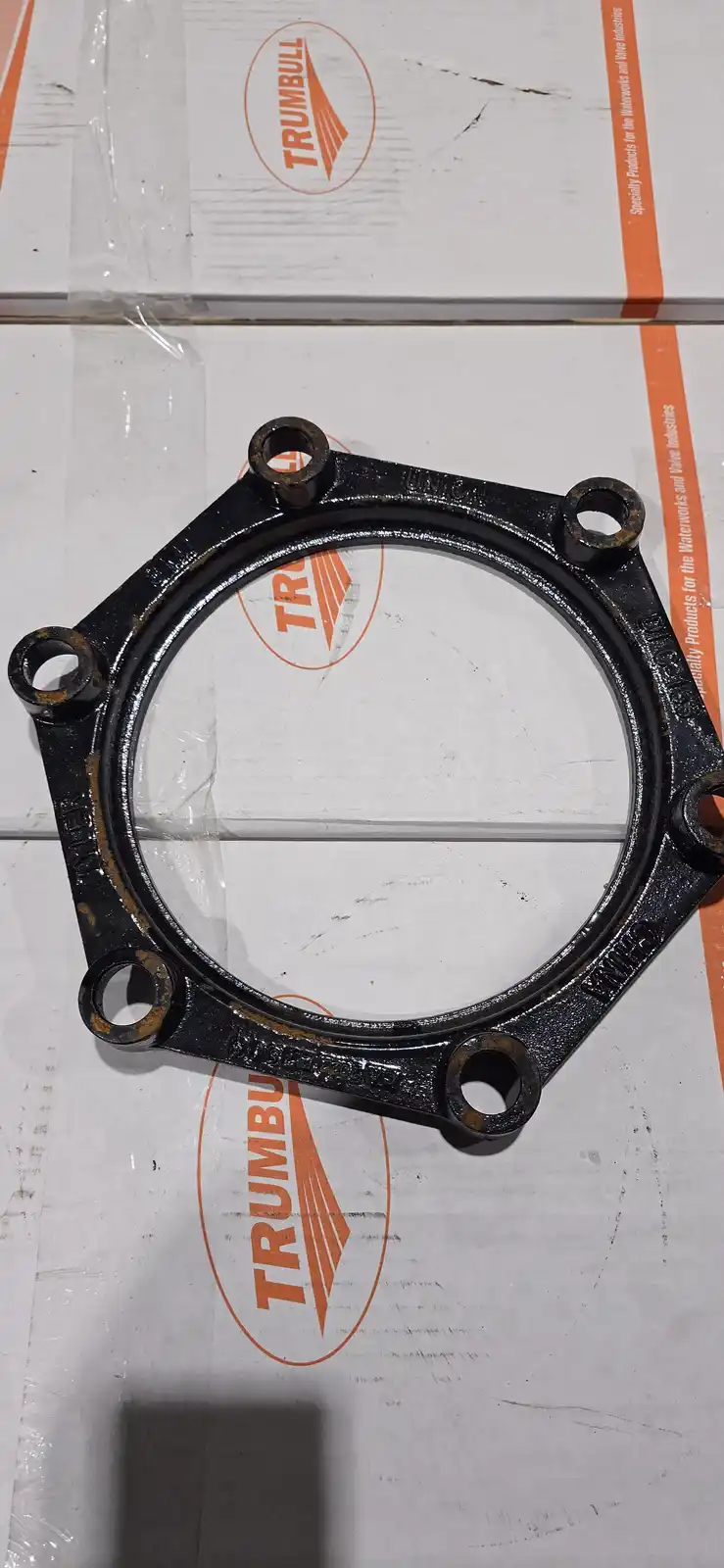 A black, circular metal pipe union with six bolt holes sits on a white cardboard box printed with the orange 'TRUMBULL' logo…