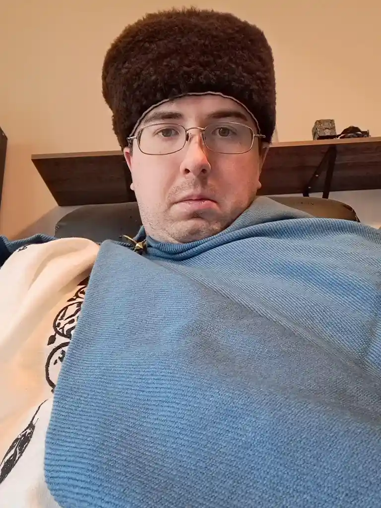 A man wearing glasses and a large, fluffy brown winter hat looks directly at the camera with a neutral expression while…