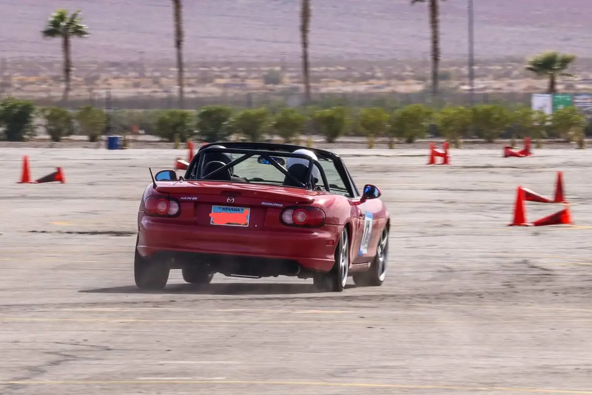 A red Mazda Miata convertible is drifting around orange traffic cones on a paved lot, kicking up dust. The car has a black…