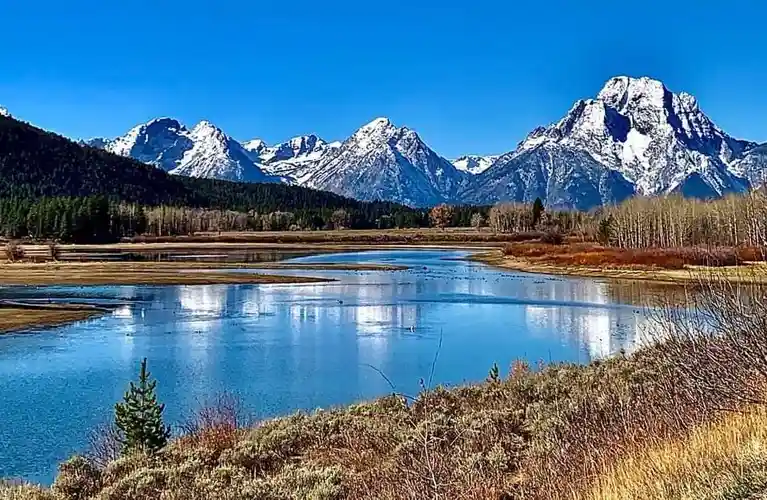 A scenic landscape photo shows a calm river reflecting snow-capped mountains under a clear blue sky, with autumnal trees and…