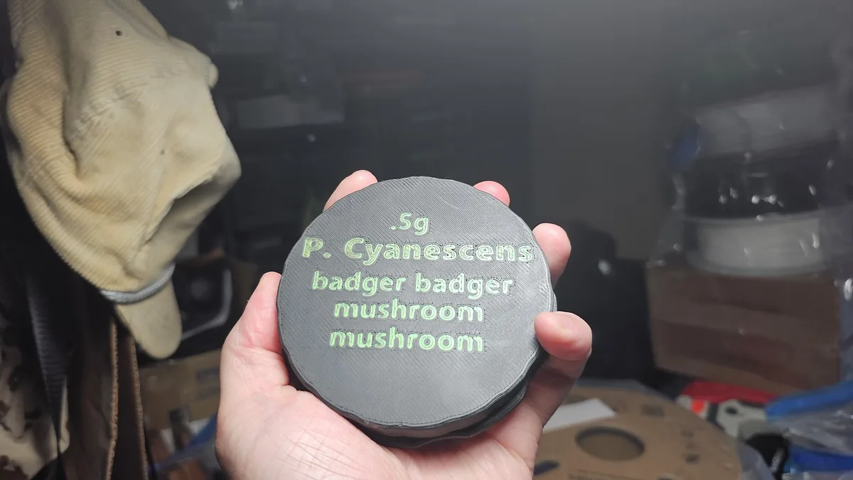 A hand holds a gray, circular, 3D-printed object with green text that reads '.5g P. Cyanescens badger badger mushroom…