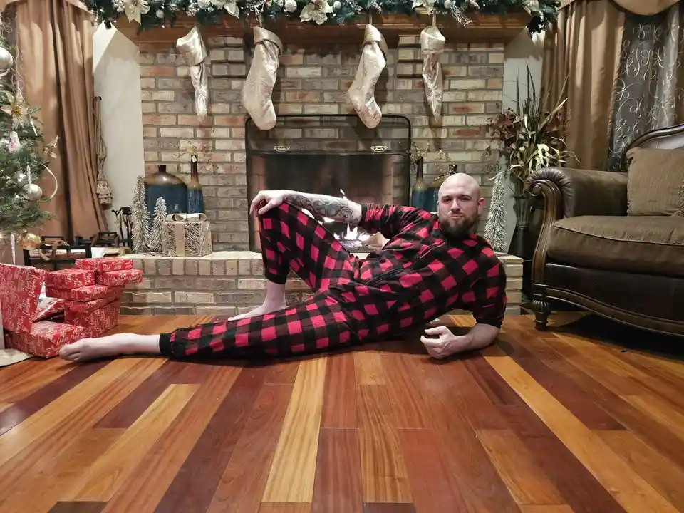 A bald man with a beard wearing red and black plaid pajamas lies on a hardwood floor in front of a decorated fireplace with…