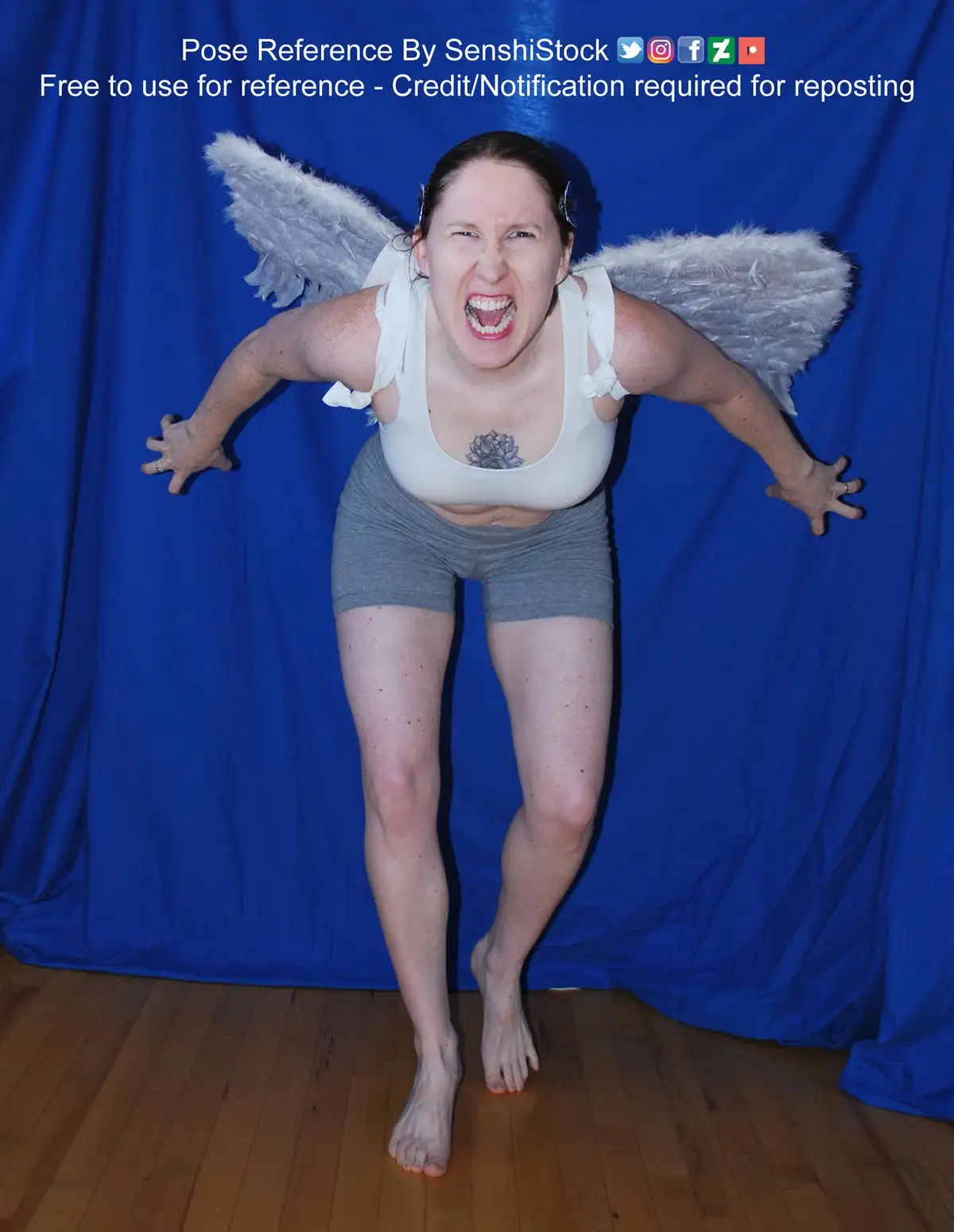 A woman wearing white angel wings and a white tank top stands in front of a blue backdrop, arms outstretched and face…