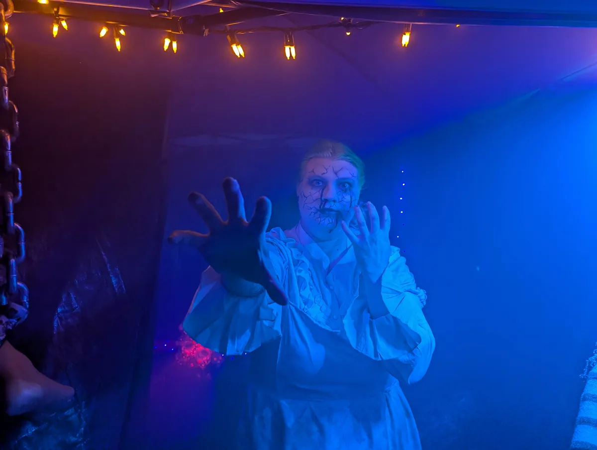 A person in a white, ruffled costume with cracked-face makeup and wide eyes, reaching out with both hands in a spooky pose…