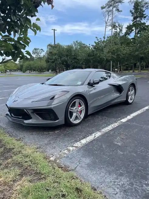 A sleek gray Chevrolet Corvette sports car is parked in an asphalt lot with white parking lines, surrounded by green trees…