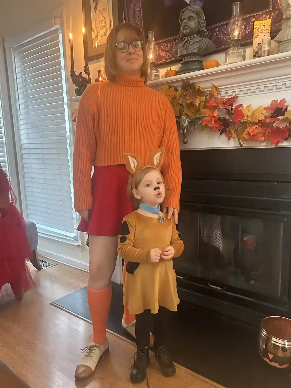 A woman and a young child pose together in front of a fireplace decorated with autumn leaves and Halloween-themed items. The…