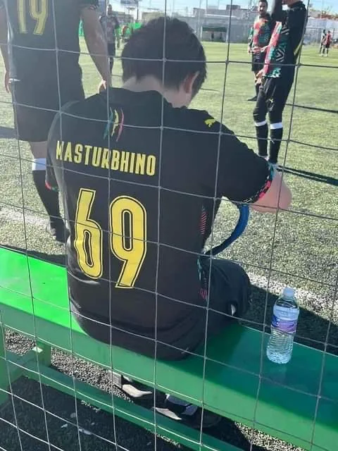 A soccer player wearing a black jersey with 'NASTURBHINO' and the number '69' in yellow is sitting on a green bench behind a…