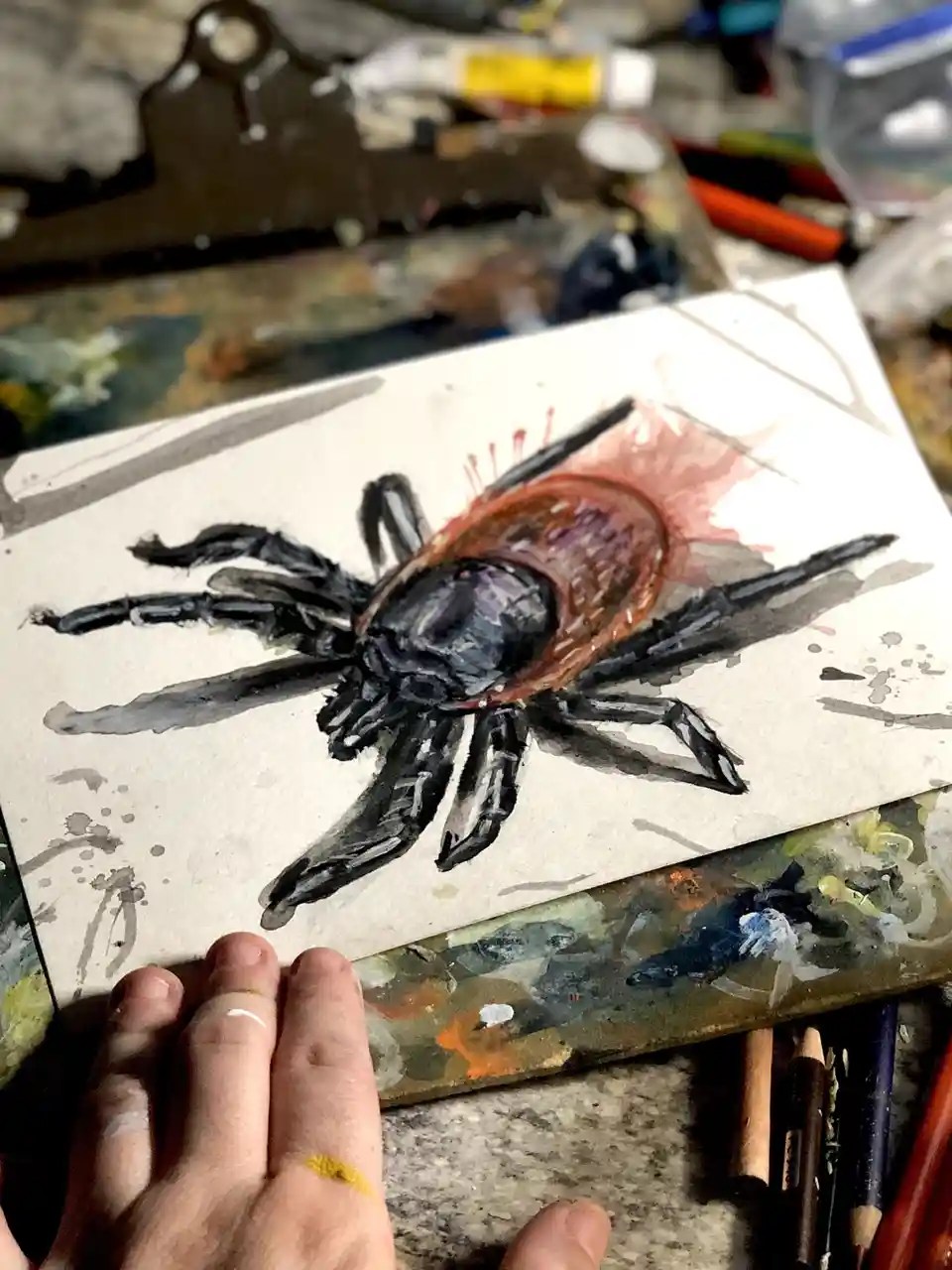 A watercolor painting of a tick with a bloated, blood-filled abdomen is shown on paper, held by an artist's hand with paint…