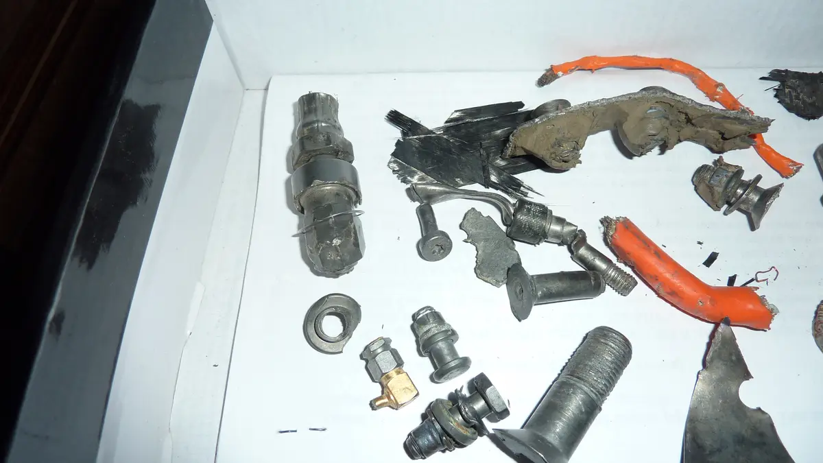 A white box contains various melted, burnt, and broken car parts including orange wires, metal bolts, and shattered…