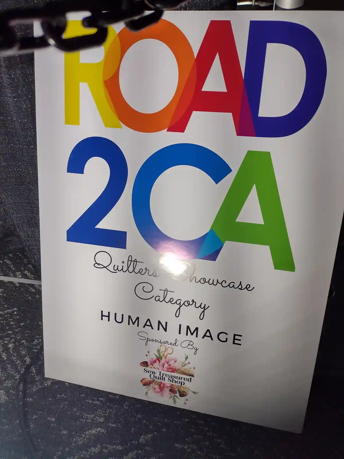A colorful sign for a 'ROAD 2CA Quilters Showcase' event, specifically for the 'HUMAN IMAGE' category, sponsored by 'Sew…