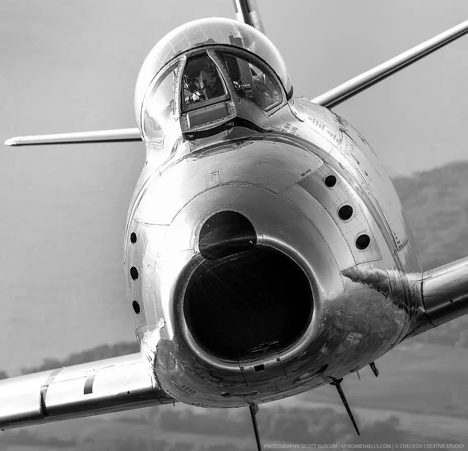 A black-and-white photo shows a close-up of a vintage jet fighter's nose, cockpit, and engine intake, with a pilot visible…