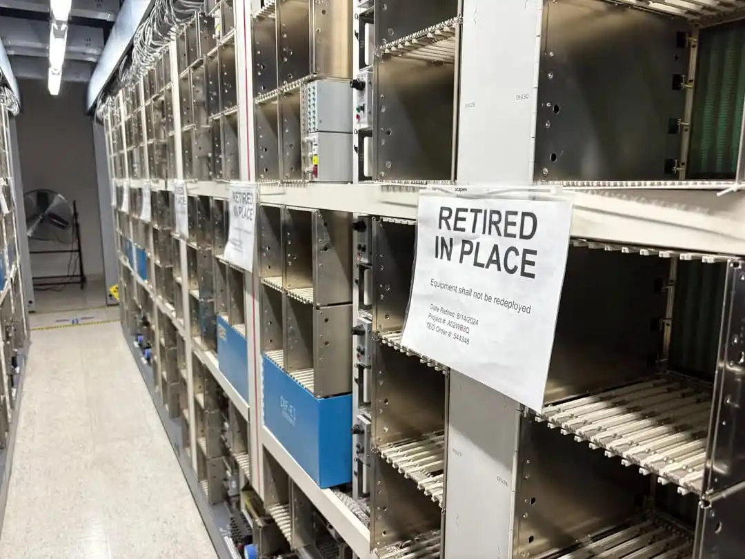 Rows of empty server racks in a data center with a prominent white sign taped to one rack that reads 'RETIRED IN PLACE' with…