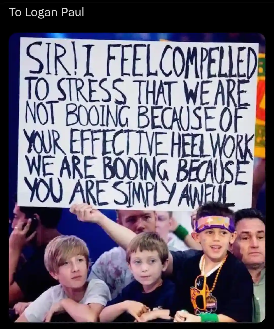 A photo of several young boys in an audience holding a large handmade sign. The sign, written in bold black marker, reads…