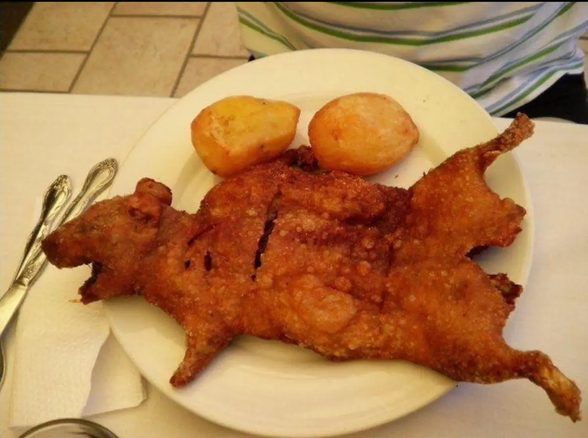 a plate of fried food shaped like a rat with two roasted potatoes beside it, served with silverware on a table, overlaid…
