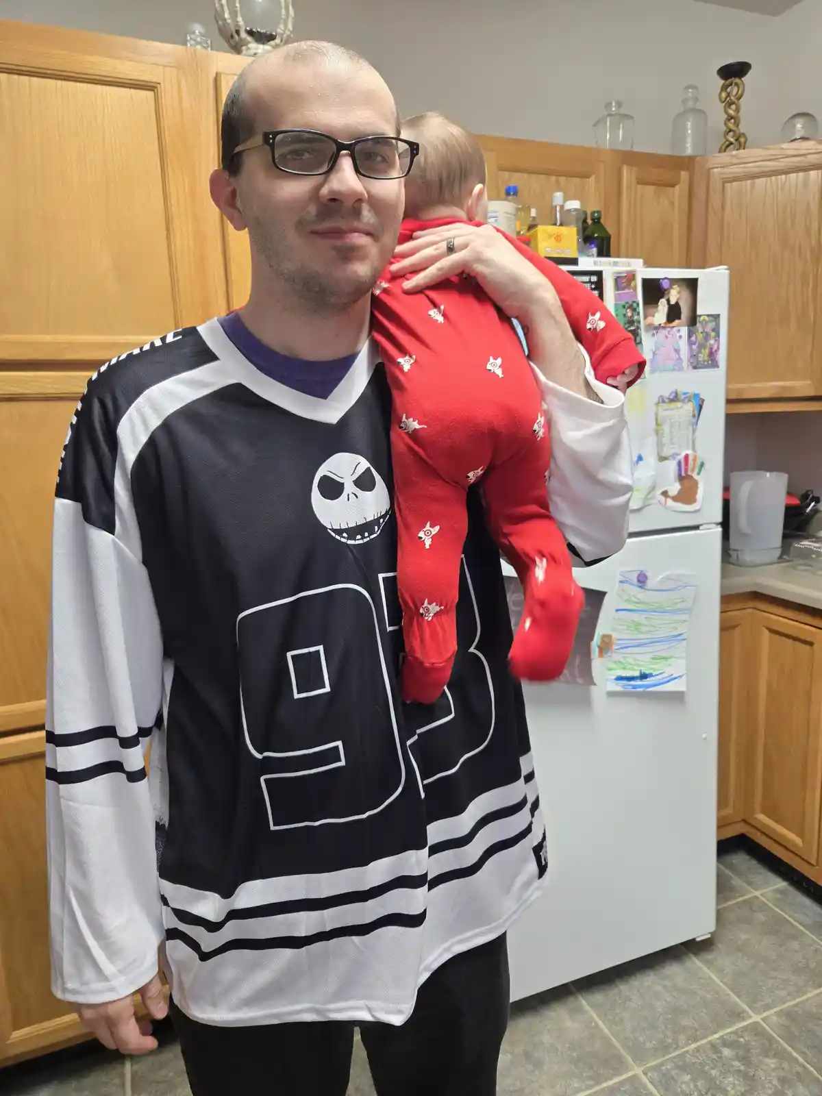 A man in a black and white hockey jersey featuring Jack Skellington's face and the number 93 holds a baby in a red onesie…