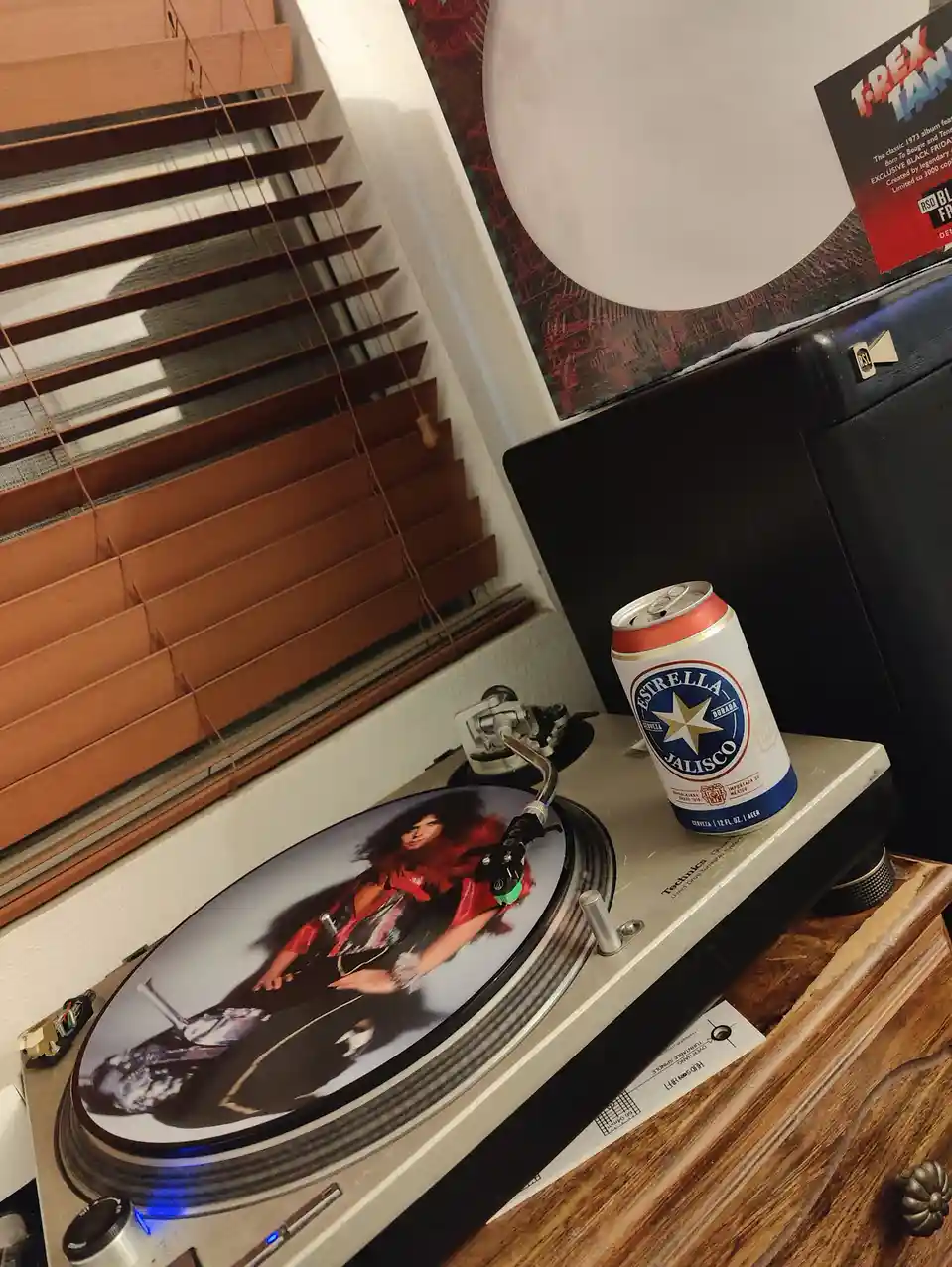 A Technics turntable sits on a wooden dresser with a beer can resting on its plinth. The turntable's platter holds a vinyl…