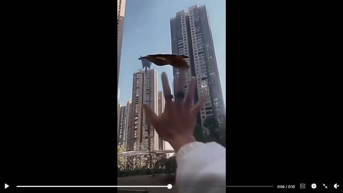 A person's hand is extended toward a large, brown, leaf-like object floating in the air against a backdrop of tall city…