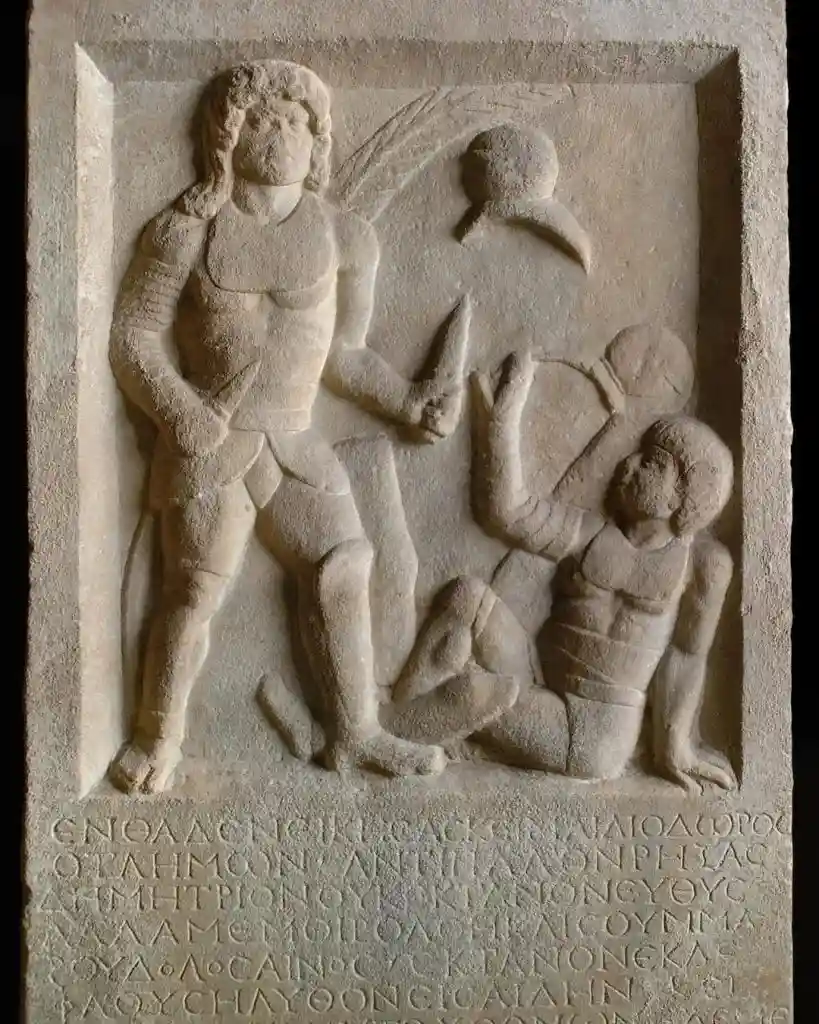 A stone relief depicts a standing warrior holding a sword over a fallen opponent, with Greek text carved at the bottom. The…