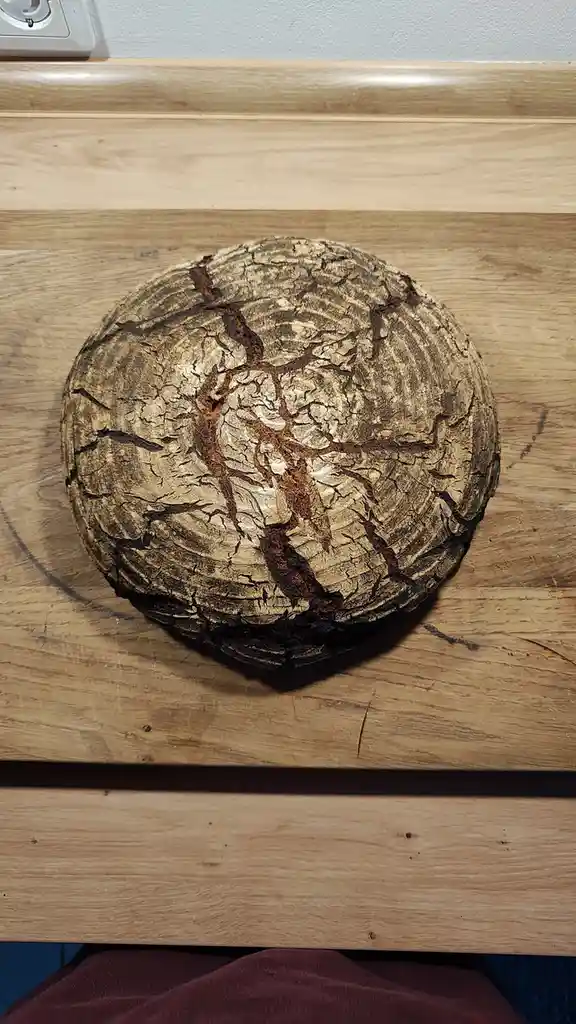 A round loaf of bread with a deeply cracked, rustic crust sits on a wooden cutting board, its surface resembling tree bark…