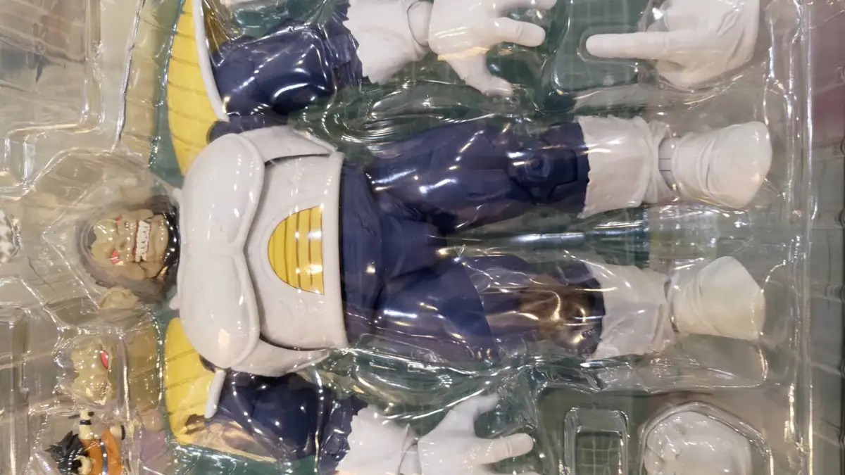 A close-up of a Dragon Ball Z action figure of Nappa, still sealed in its clear plastic packaging, lying on its back with…