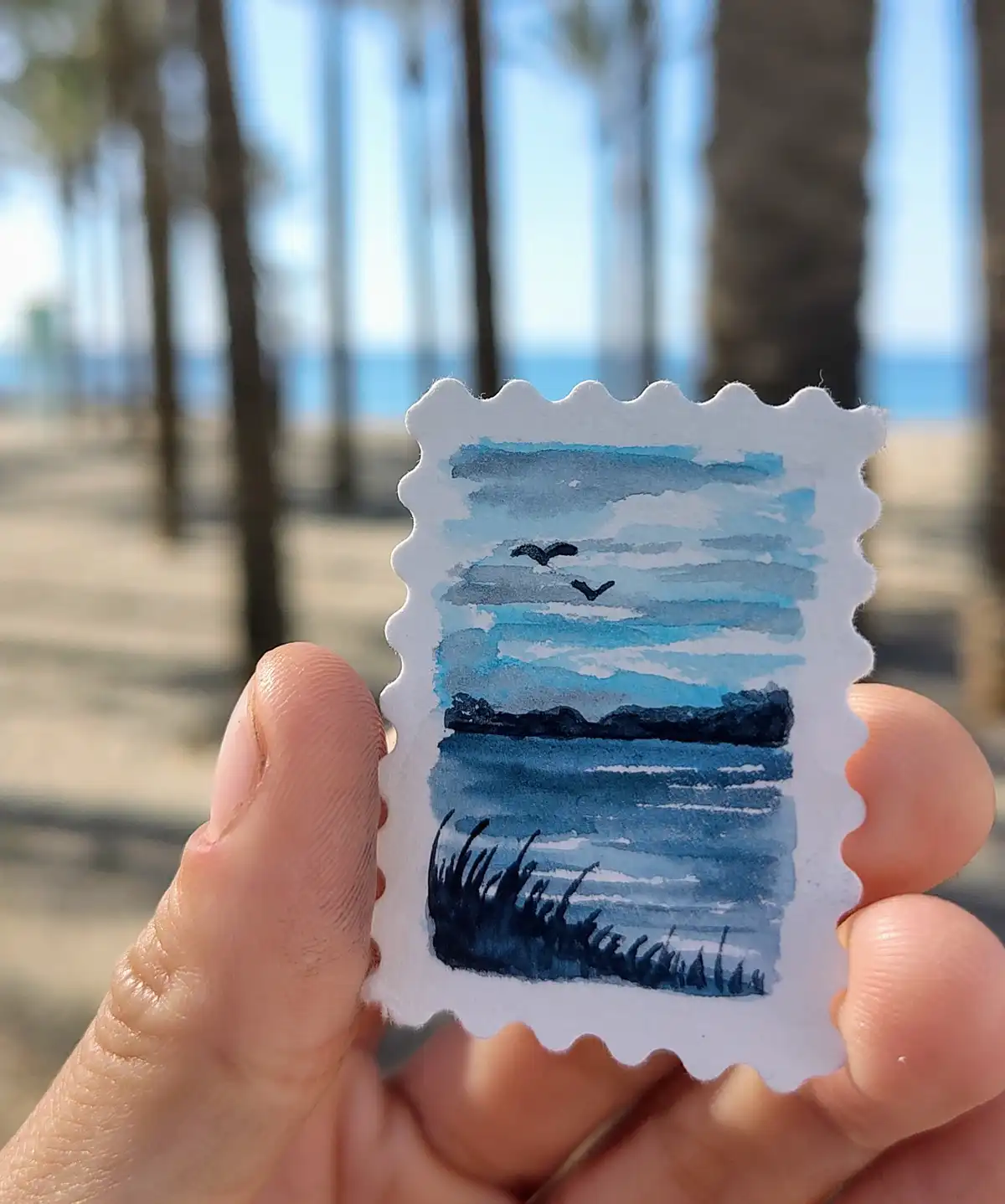 A hand holds a small, scalloped-edged watercolor painting resembling a postage stamp, depicting a coastal scene with blue…