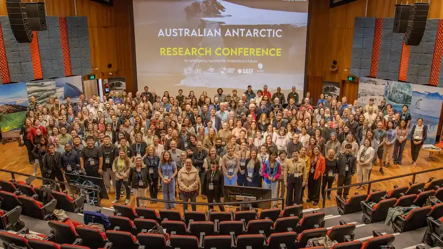 A large group of people in a conference hall pose for a group photo in front of a large screen displaying 'AUSTRALIAN…