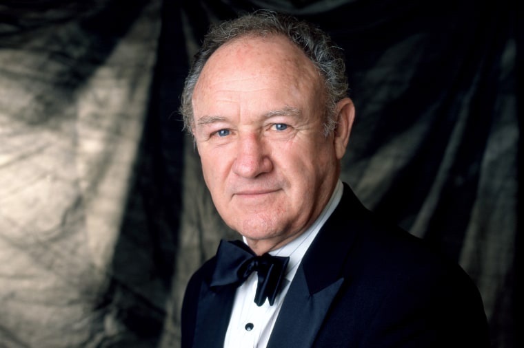 a formal portrait of a man with gray hair and blue eyes wearing a black tuxedo and bow tie against a dark draped background…