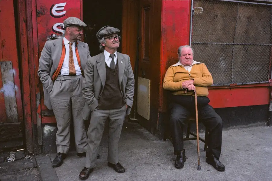 Three older men stand or sit outside a red building; two lean against a doorway wearing suits and flat caps, while the third…