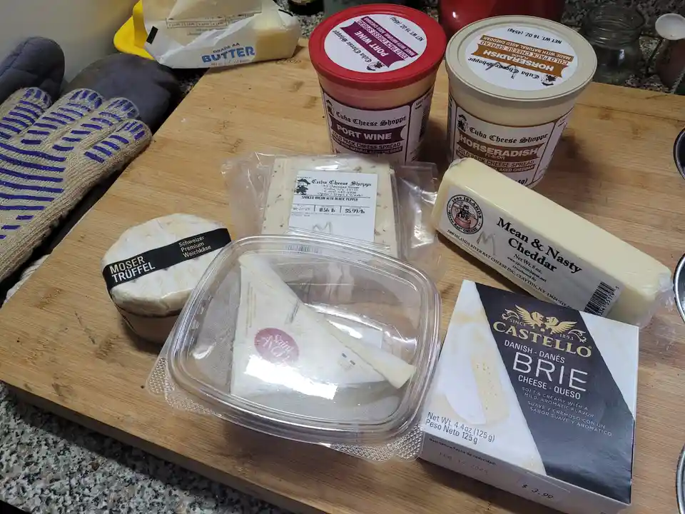 A wooden cutting board displays an assortment of expensive and varied cheeses and dairy products, including a block of 'Mean…