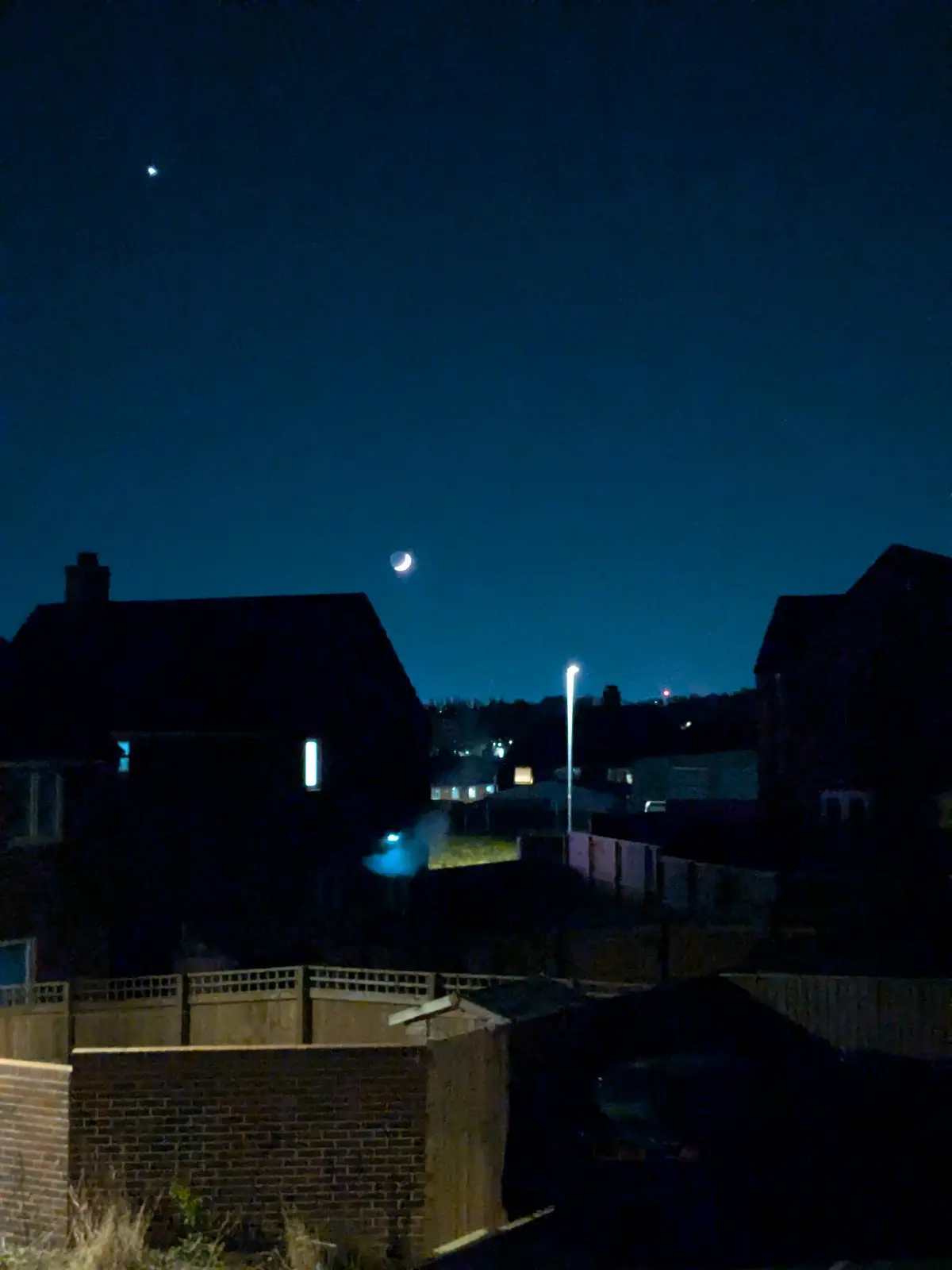 A nighttime photo of a residential neighborhood with houses, fences, and a streetlamp under a dark blue sky. A crescent moon…