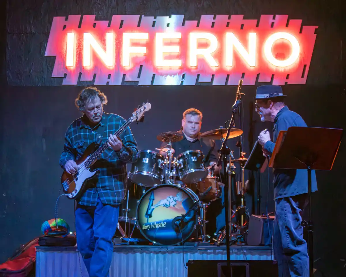 A band performs on stage under a large, glowing red neon sign that reads 'INFERNO'. The bassist on the left wears a plaid…