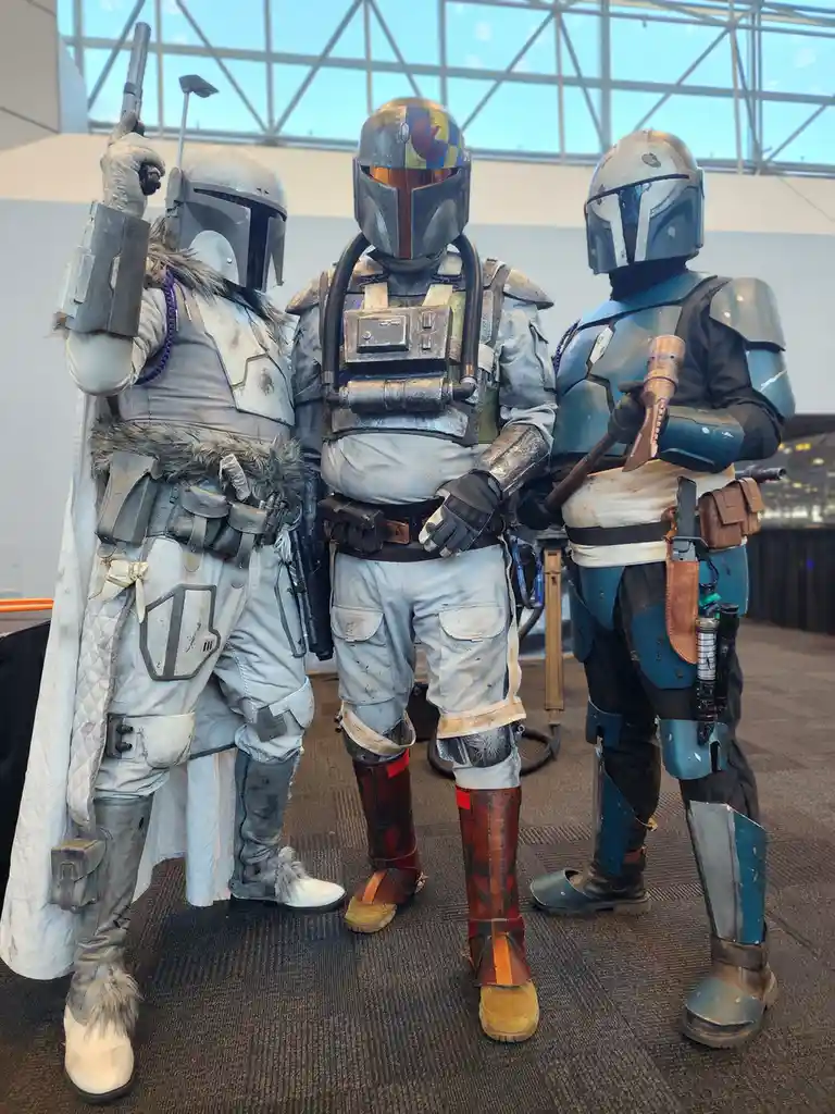 Three people in detailed Mandalorian armor cosplay stand side-by-side indoors, each with unique helmet designs and gear. The…