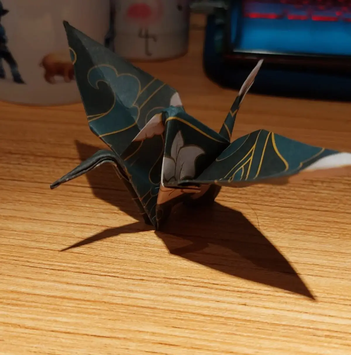 A dark green origami crane with gold and white floral patterns sits on a wooden desk, casting a sharp shadow. In the…