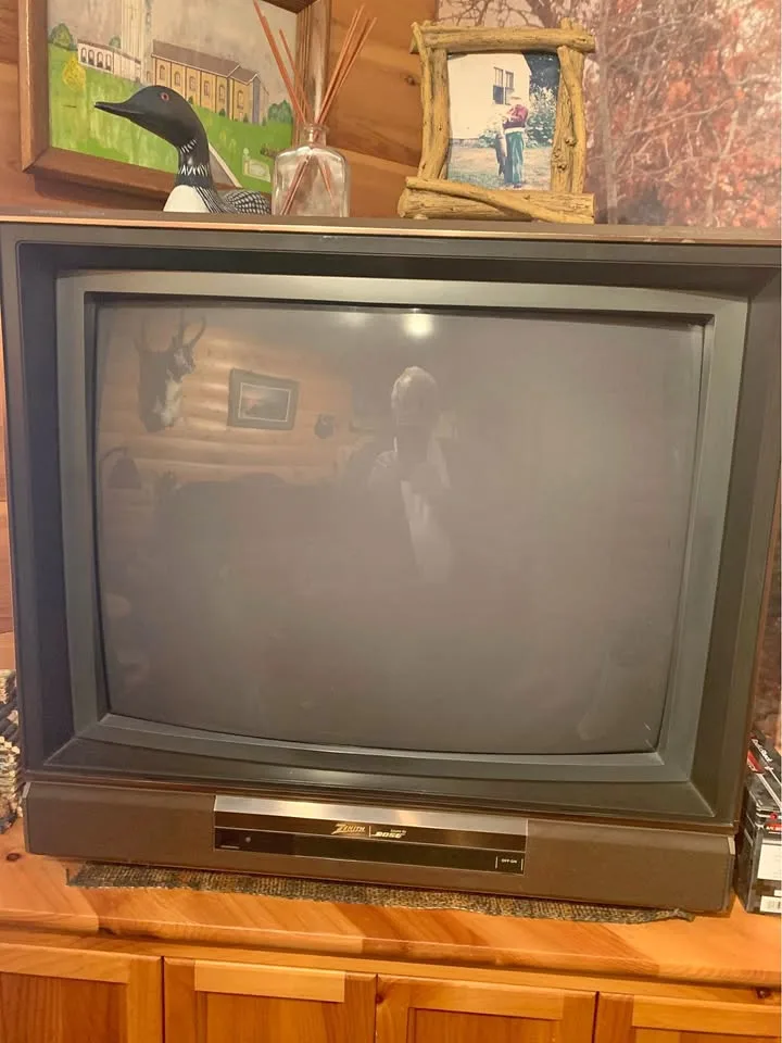 A person is taking a selfie in the reflection of an old, dark, CRT television screen. The TV sits on a wooden cabinet, with…