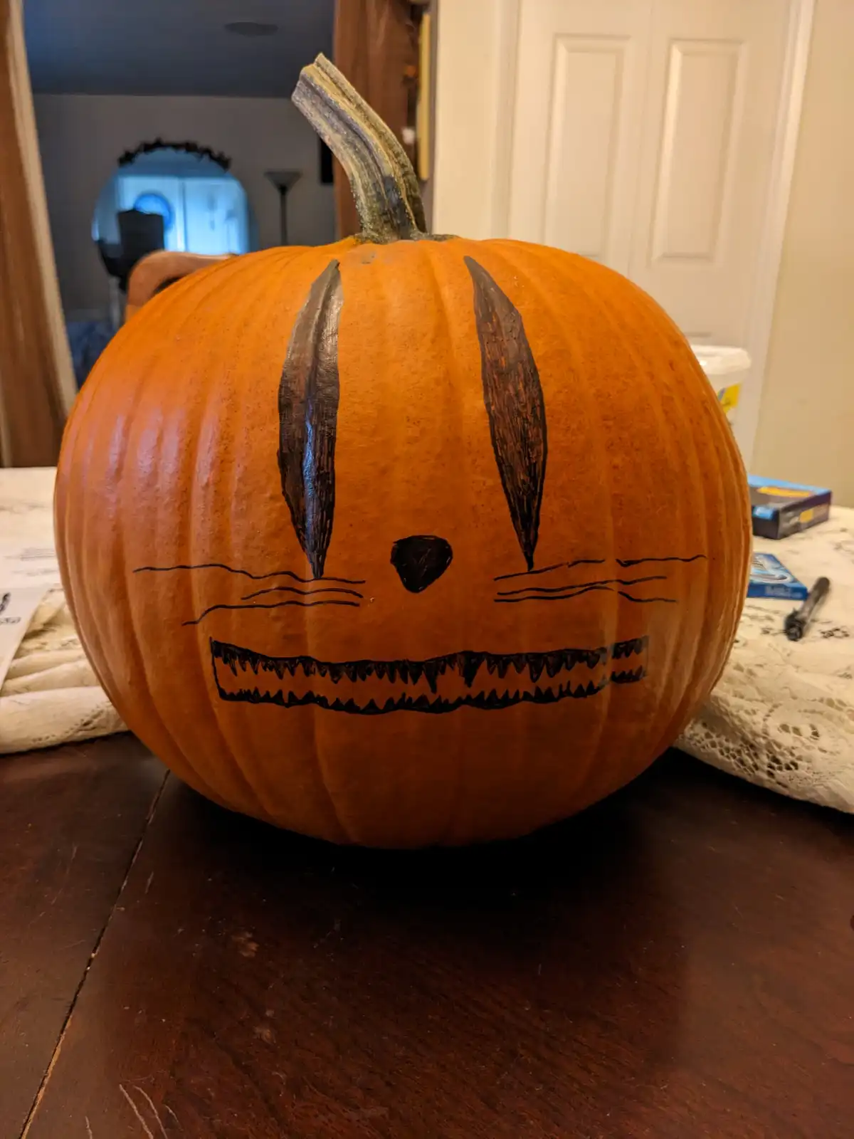 An orange pumpkin sits on a wooden table, decorated with black marker to resemble a bunny face with long ears, whiskers, and…