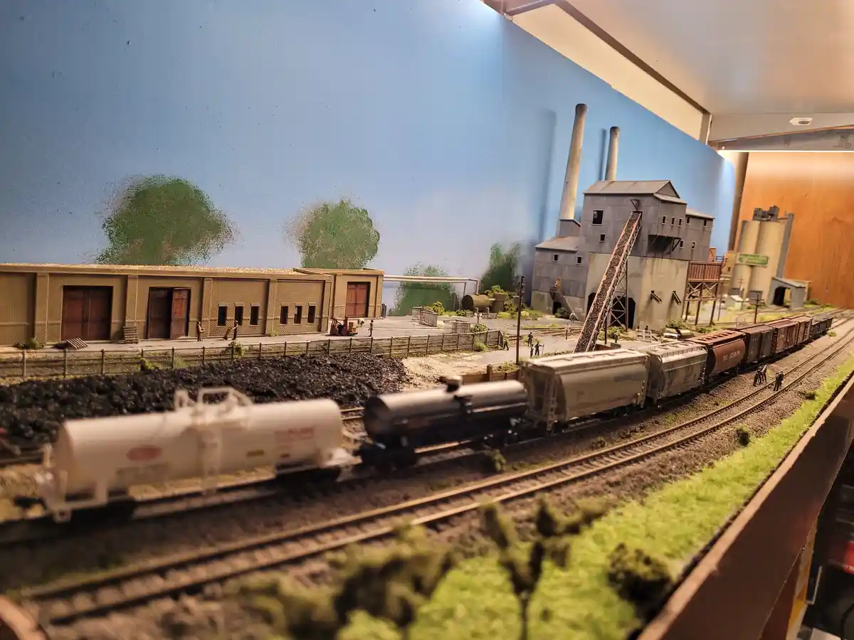 A detailed model train layout featuring miniature trains, industrial buildings, and tiny figures, with overlaid text reading…