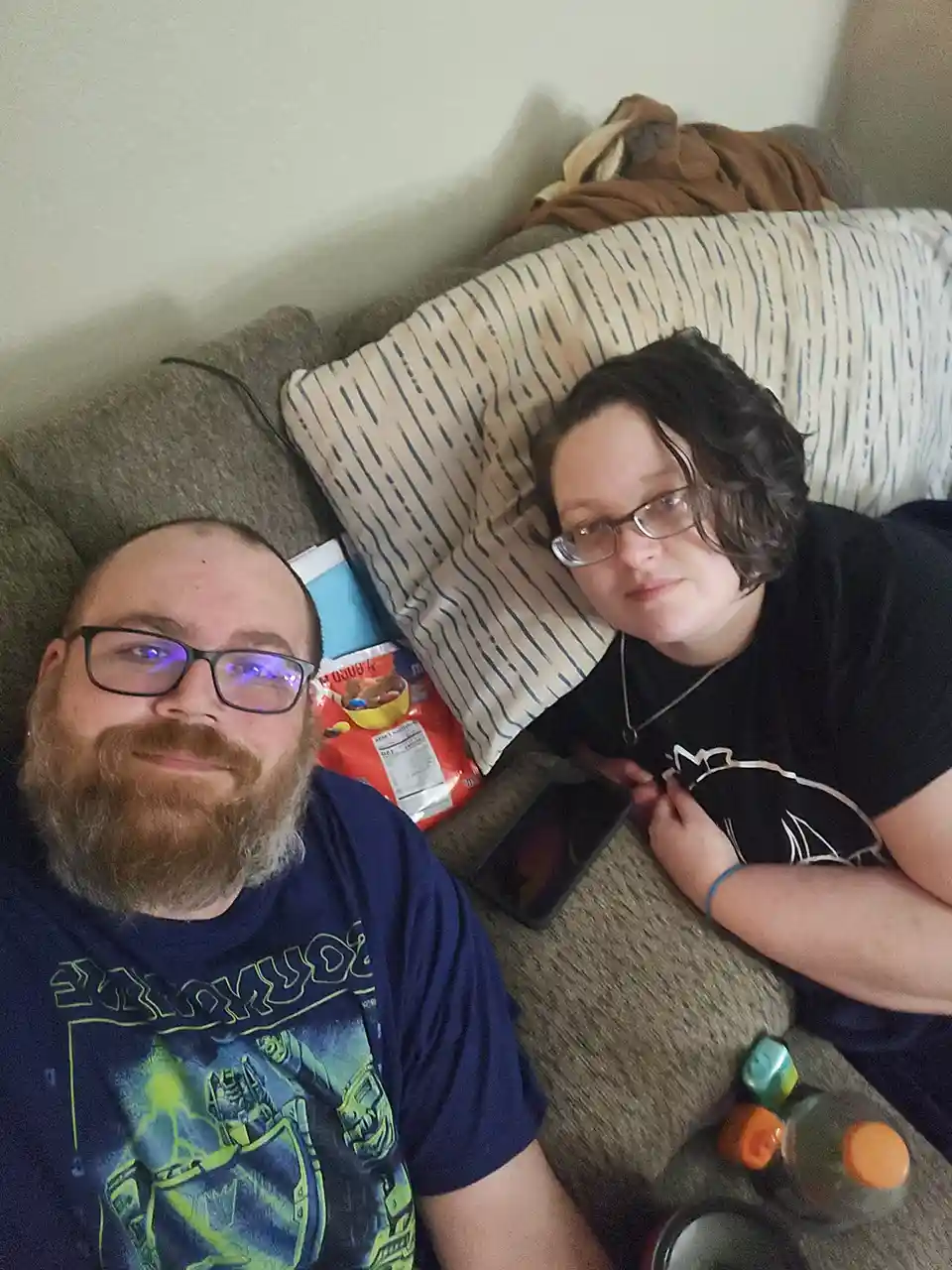 Two people are lying on a couch; the person on the left smiles at the camera while the person on the right has a bored or…