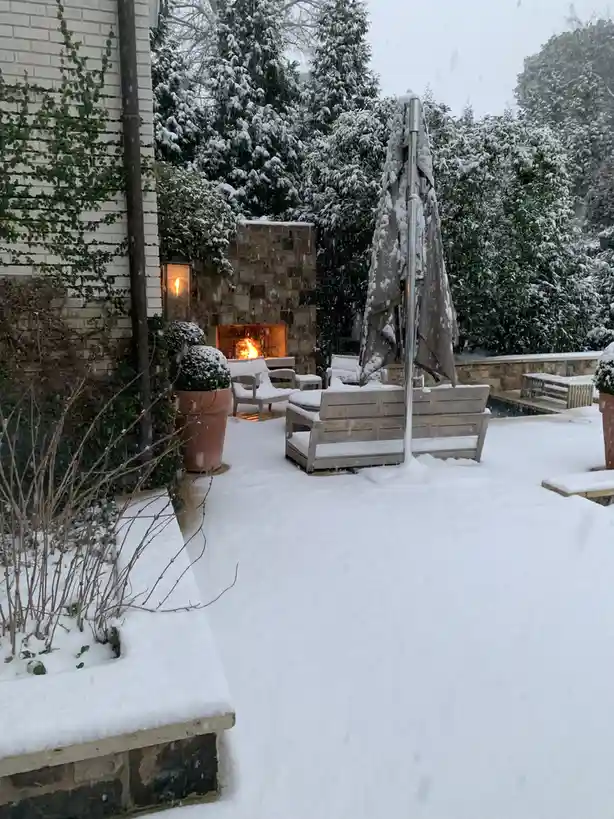 A snowy backyard patio features snow-covered outdoor furniture, including a closed umbrella and lounge chairs, with a stone…