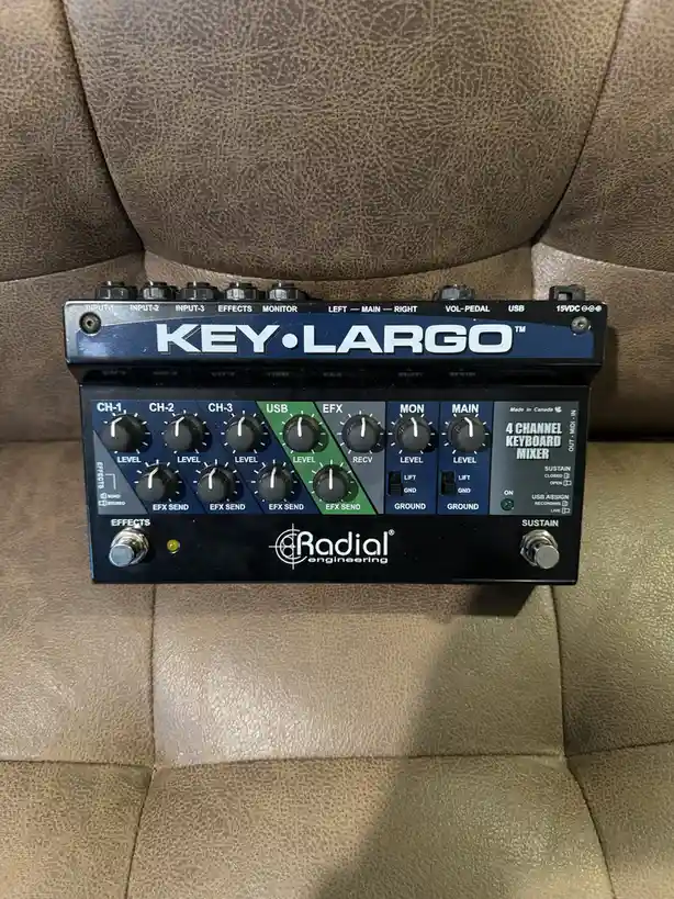 A Radial Engineering Key-Largo 4-channel keyboard mixer sits on a brown leather couch, its knobs and labels clearly visible…