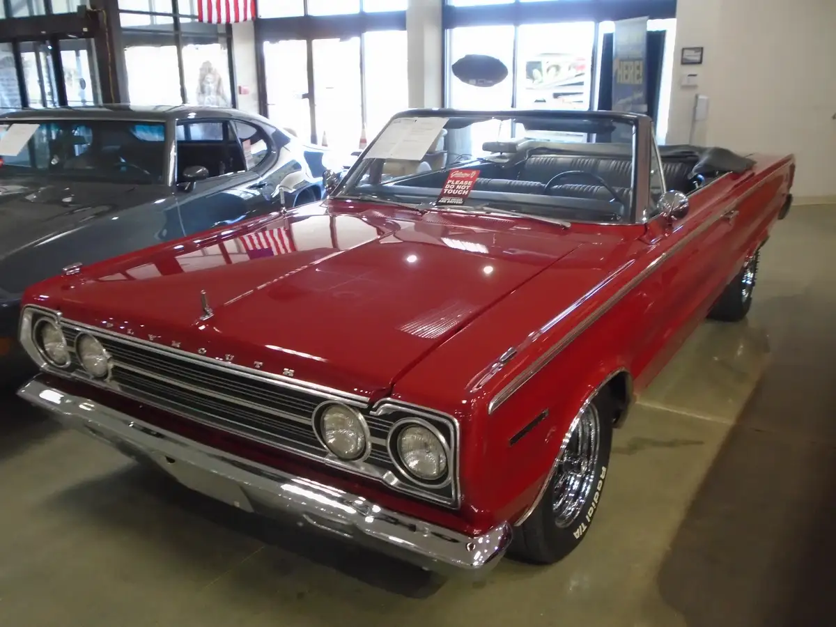 A shiny red 1960s Plymouth convertible is displayed indoors, likely in a museum or dealership, with a 'PLEASE DO NOT TOUCH'…
