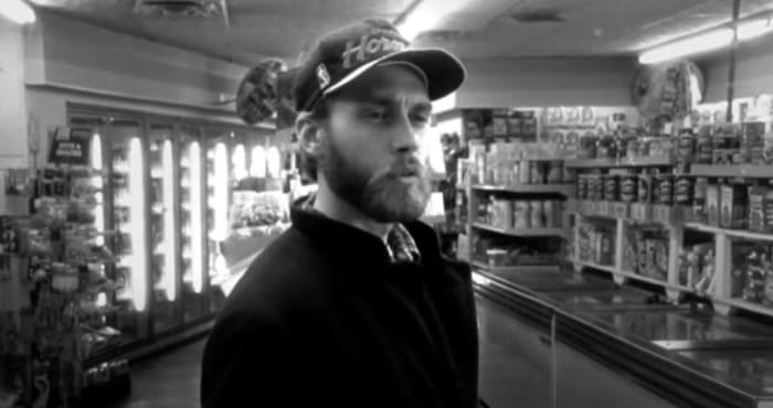 A black-and-white image of a man with a beard and a baseball cap standing in a grocery store aisle, looking off to the side…