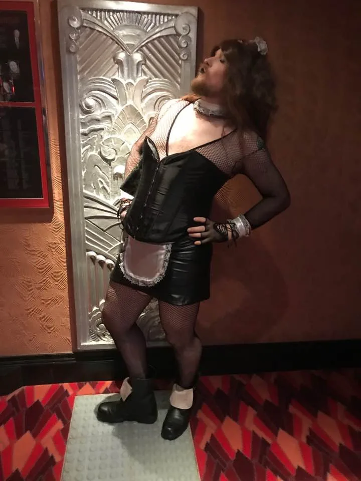 A person with a beard and long hair, wearing a black corset, fishnet sleeves, a leather mini-skirt with a white apron…