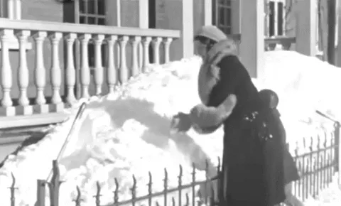 A black-and-white sequence shows a woman in a long coat and scarf building a snow mound, then dramatically throwing snow…