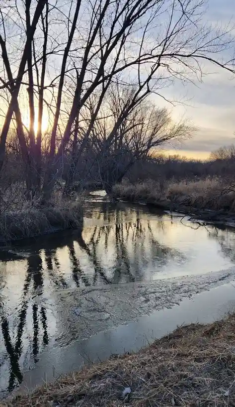 A serene winter scene shows a partially frozen river reflecting bare trees against a sunset sky, with the overlaid text…